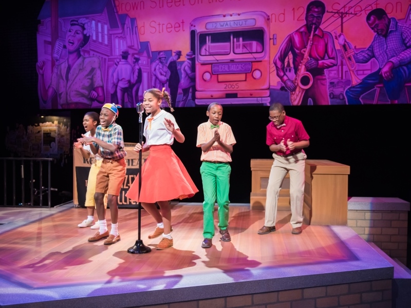 Children perform on stage against a music themed background, with colorful 1950s clothing and a vintage microphone