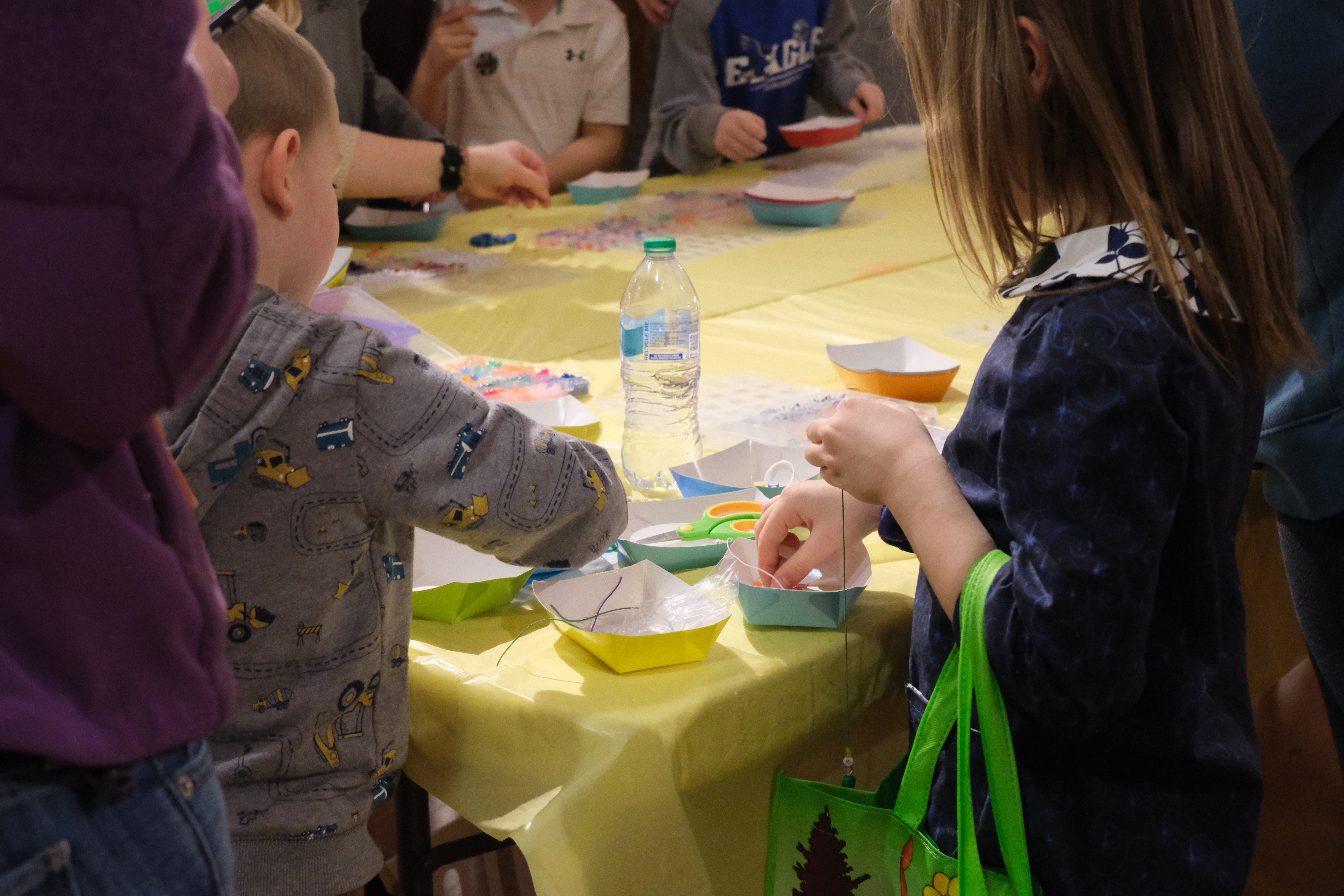 Children are gathered around a table with a yellow tablecloth, engaged in a craft activity using beads and string